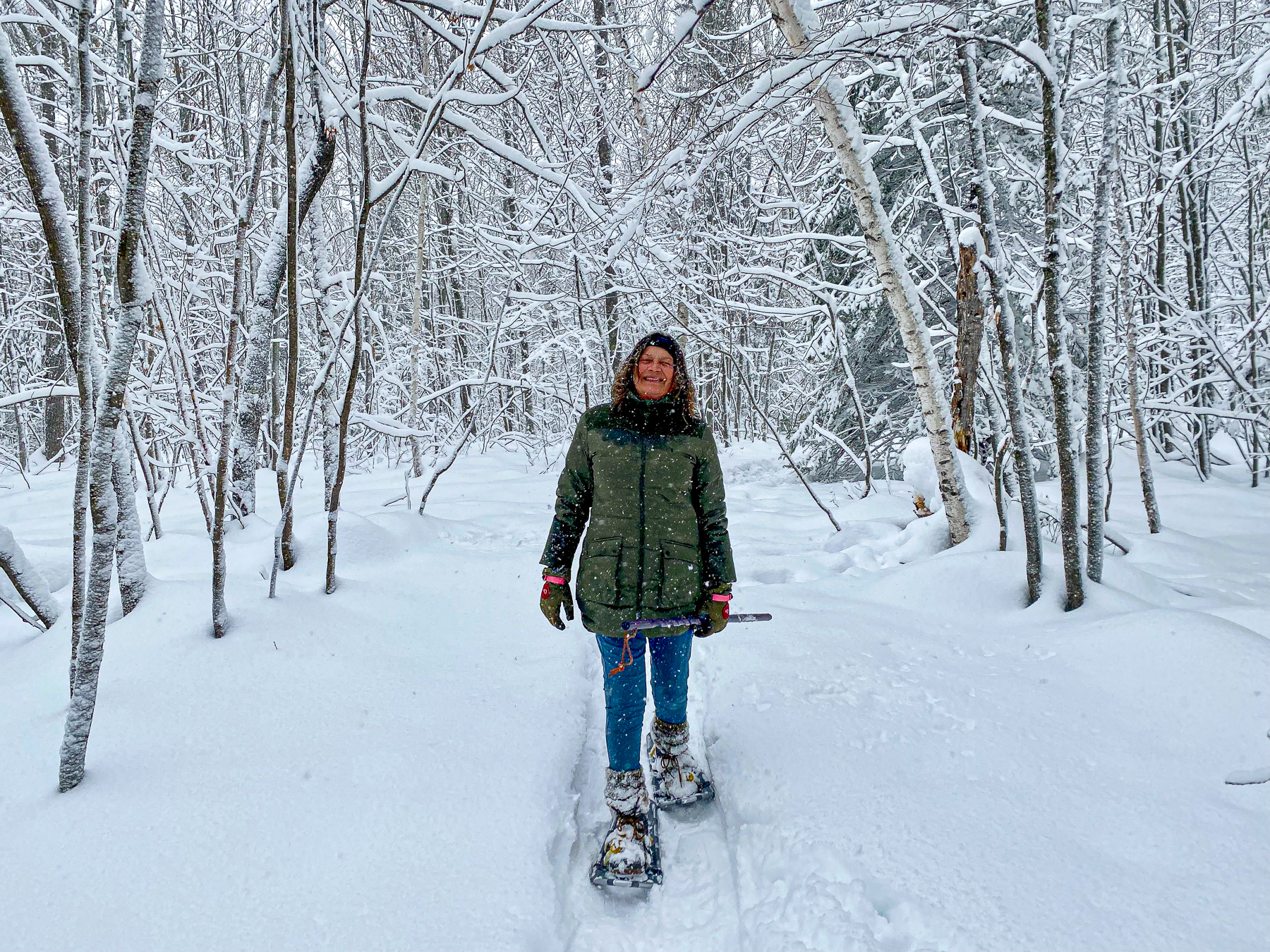 A person in winter gear cross-country skis along a snowy forest path with snow-covered branches on both sides.